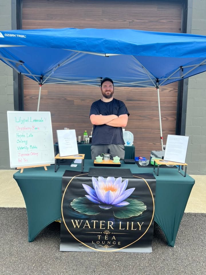 Water Lily Tea Lounge's owner standing behind the booth with the lotus banner and green tea table